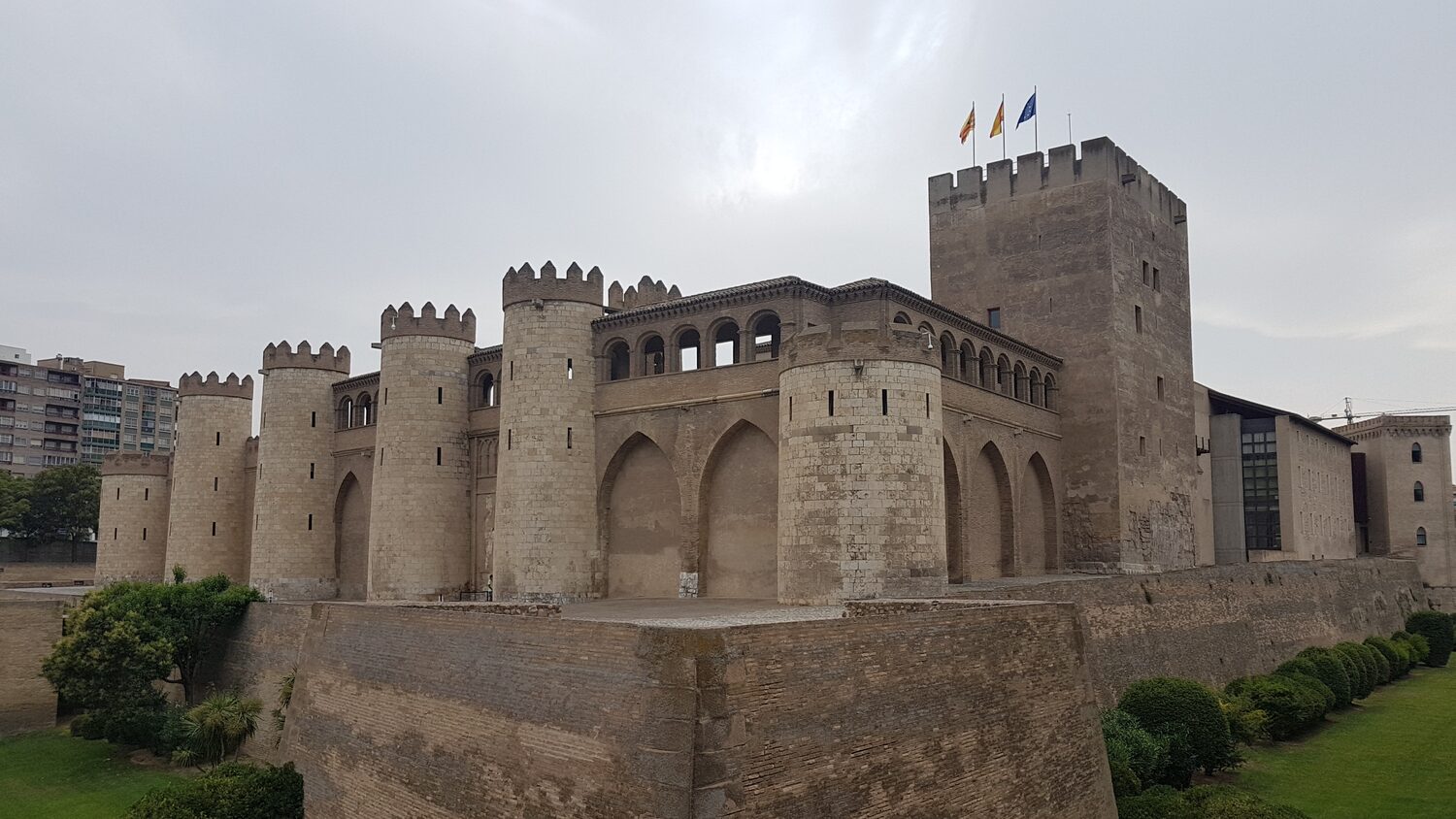 el palacio de la Aljafería de Zaragoza visto de tres cuartos, con sus torres cilíndricas almenadas y la torre del Trovador al fondo