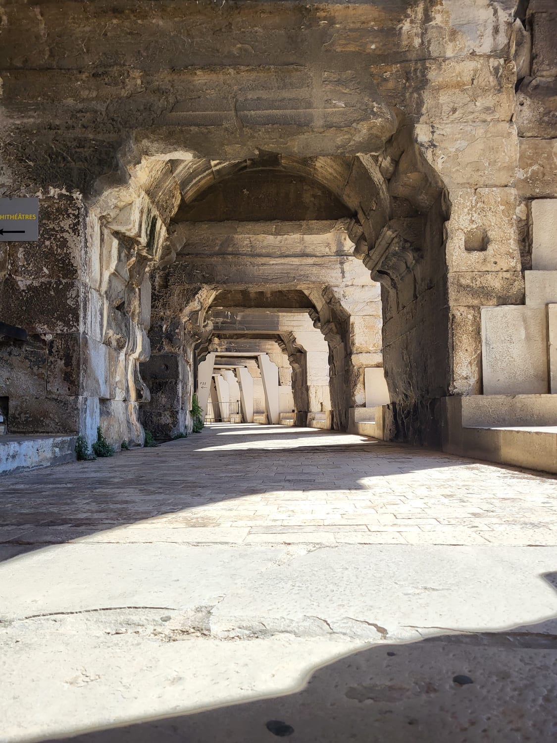 la galerie extérieure du rez-de-chaussée des Arènes de Nîmes, avec ses piliers, ses voûtes de pierre et la signalétique de l’amphithéâtre