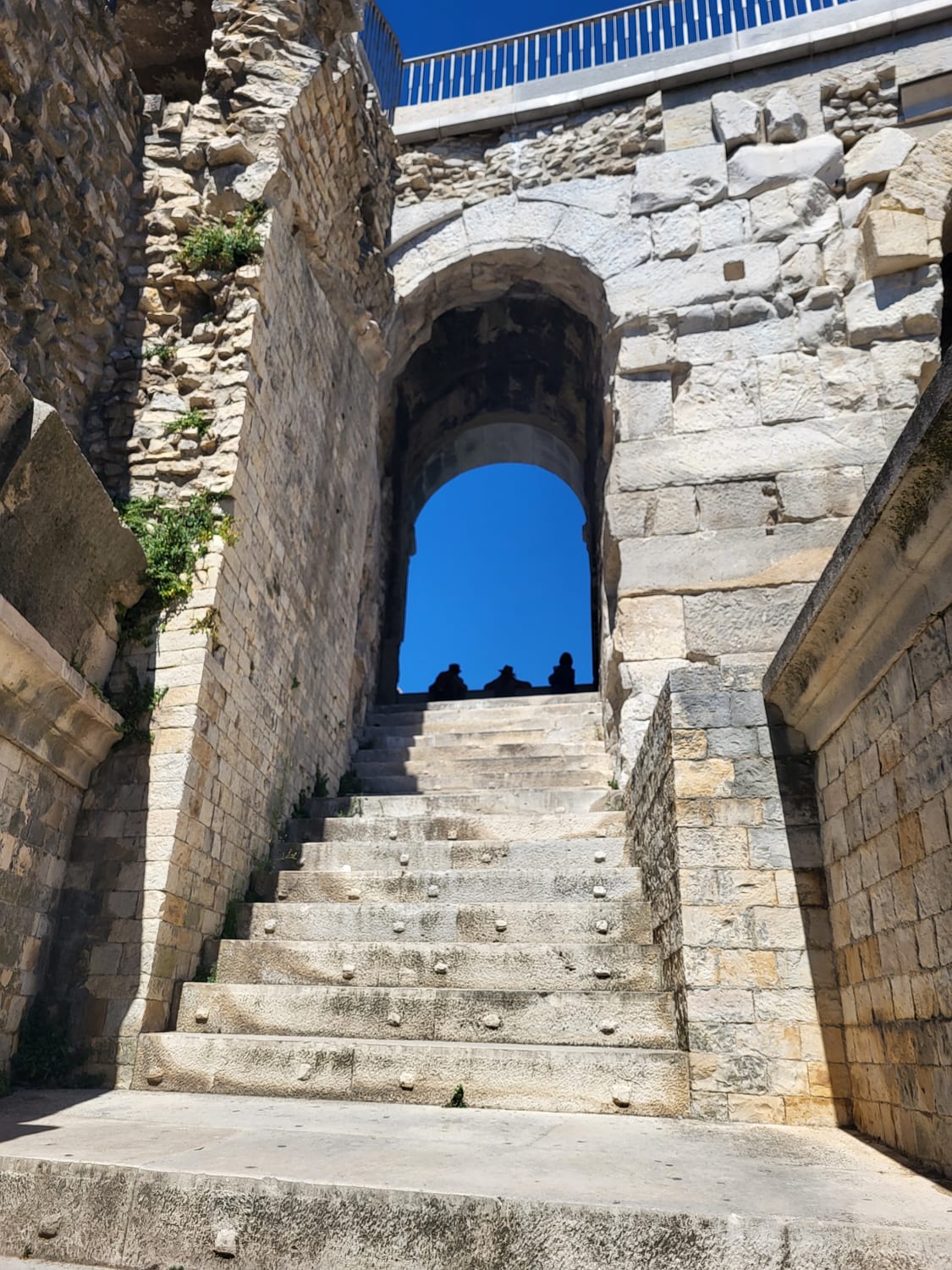 un escalier de pierre des Arènes de Nîmes montant vers l’arc d’un vomitorium, avec les silhouettes de quelques visiteurs sur les gradins