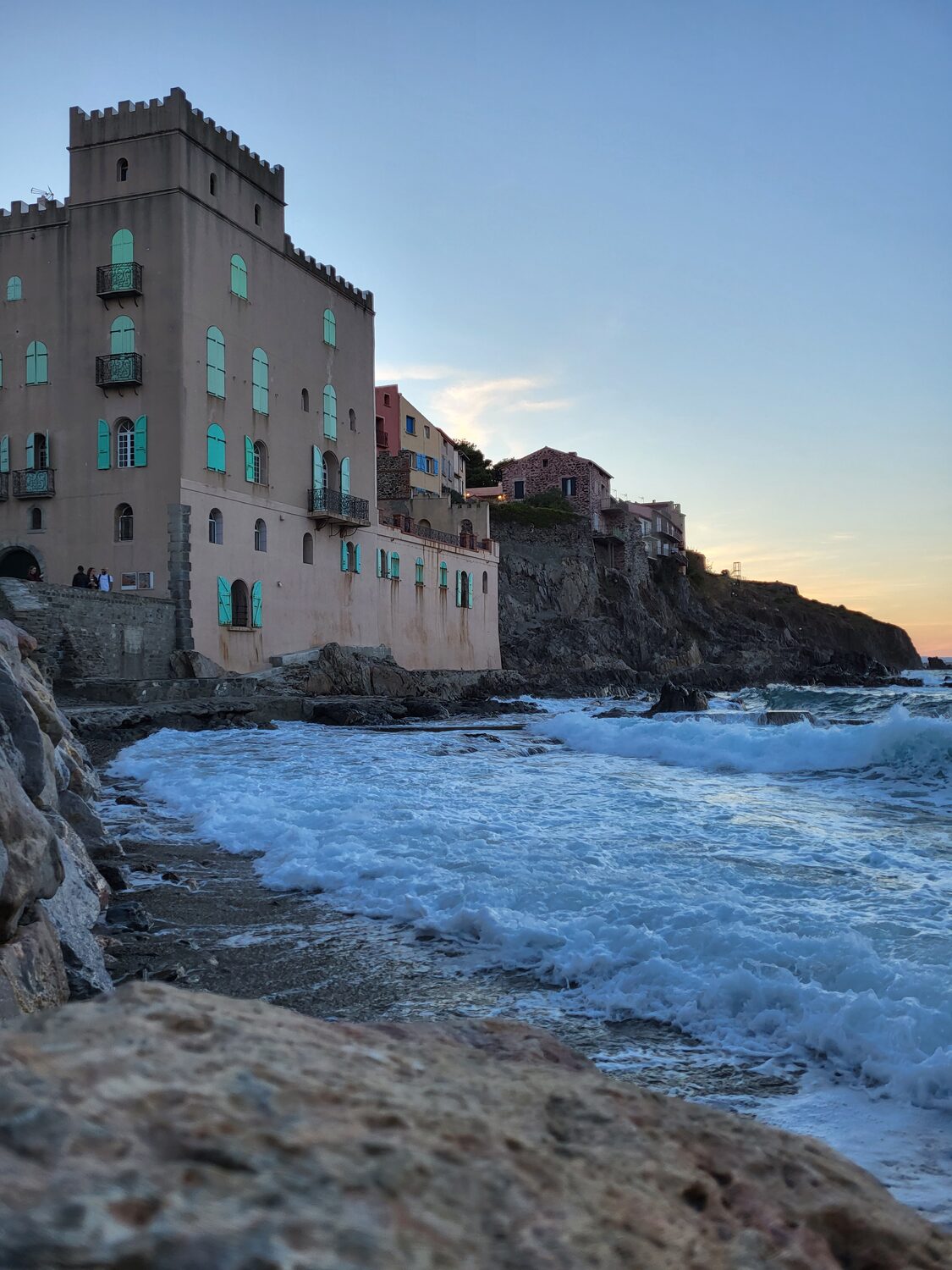 una casa merlata di Collioure sopra gli scogli, con le imposte verde acqua e le onde che frangono